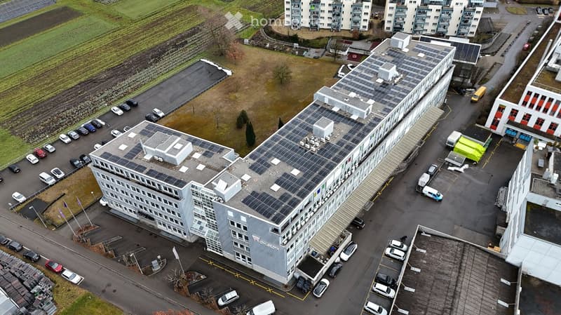 Multi-story building, gray exterior, solar panels on the roofs, many parking spaces in front