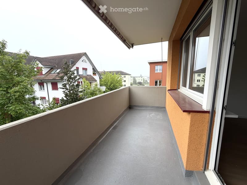 Balcony / Terrace. Balcony with grey concrete floor, beige walls, and wooden railing, overlooking a residential area with houses and trees.