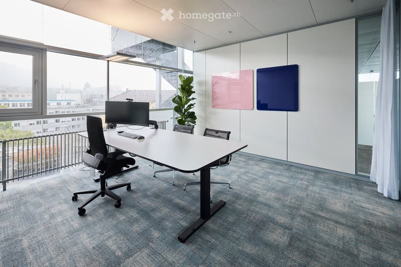 Modern office space with a large white table, ergonomic chairs, computer setup, and a potted plant near large windows offering a city view.