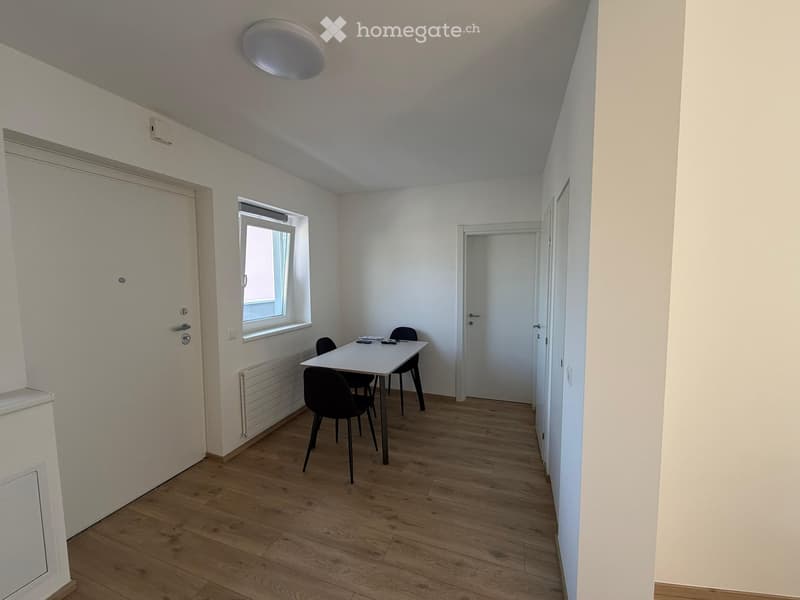 Furnished Room. A small, unfurnished room with wooden flooring, white walls, and a single window. A small table and chairs are present, along with a radiator and a closed door.