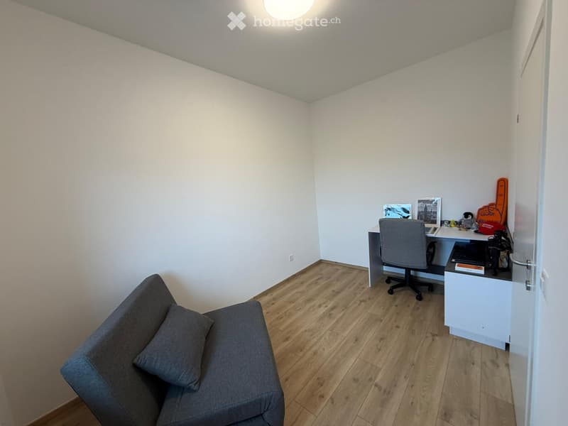 Furnished Room. A small, modern room with a grey sofa, a white desk with a chair, and various decorative items on the desk. The room features light wooden flooring and white walls.