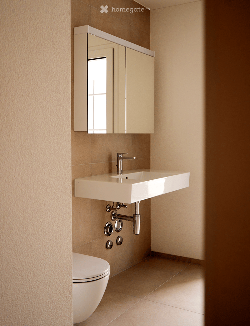 Bathroom. Modern bathroom with a white sink, chrome faucet, mirrored cabinet, and a toilet. The walls are tiled in a warm beige color, and the floor is covered with light-colored tiles.
