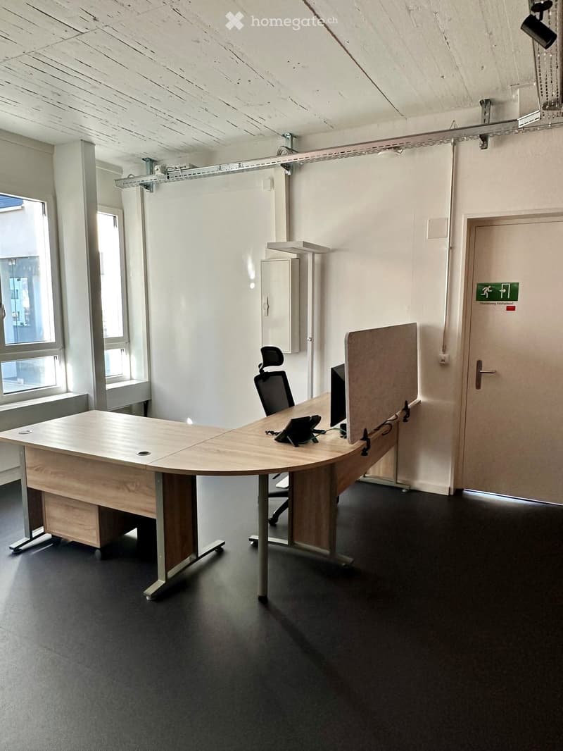Furnished Room. Modern office space with a large wooden desk, ergonomic chair, computer screen, and screen partition. The room features white walls, large windows, and a dark grey carpeted floor.
