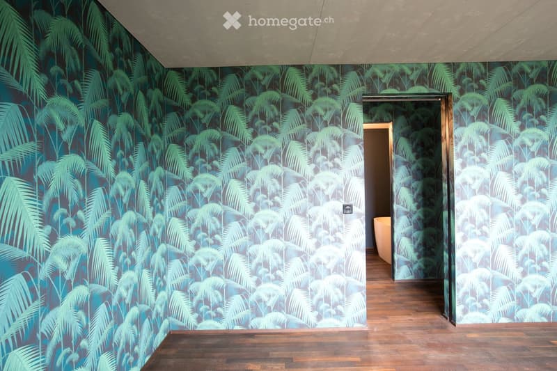 Empty room with wooden flooring and tropical-themed wallpaper featuring palm leaves. A doorway leads to another room with a visible bathtub.
