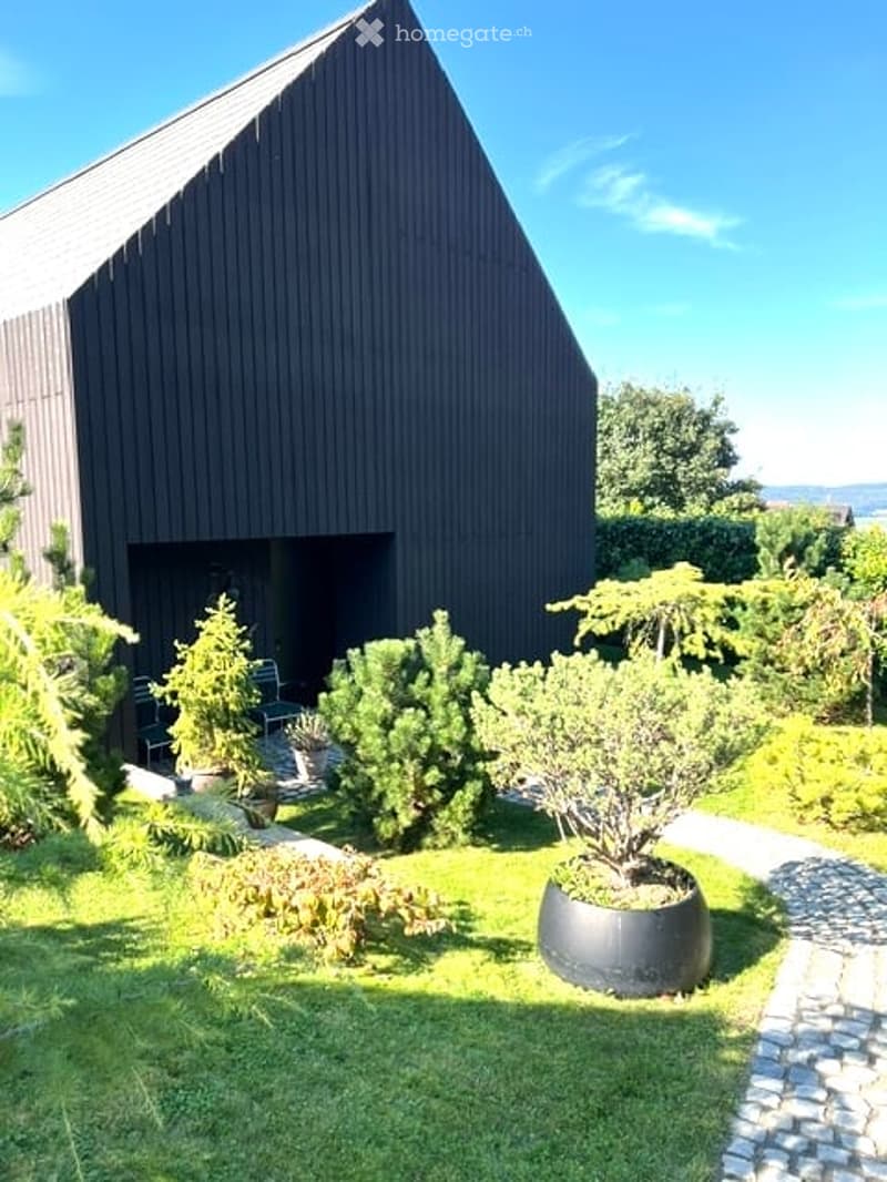 Modern black house with a sloped roof, surrounded by a well-maintained garden with various potted plants and shrubs. The garden features a gravel pathway leading to the house entrance.
