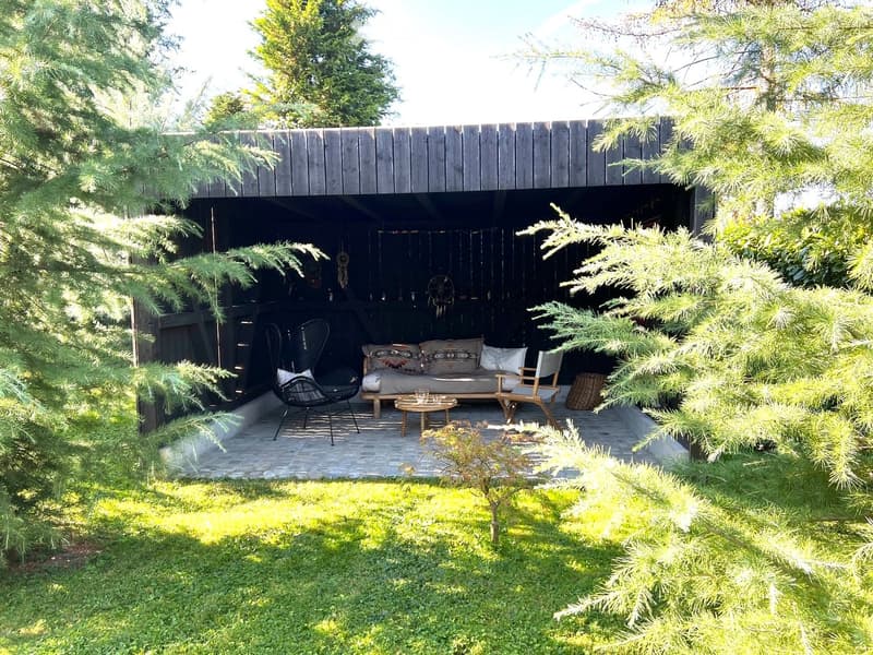 A cozy outdoor seating area with a wooden structure, featuring a sofa, a small table, and two chairs, surrounded by greenery and trees.