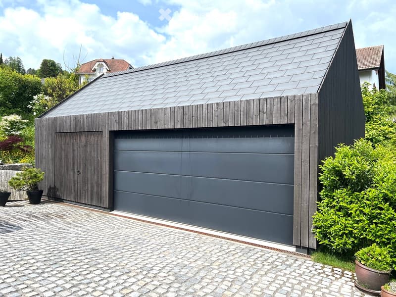 Modern, single-story garage with dark wooden exterior and grey garage door, surrounded by greenery and potted plants.