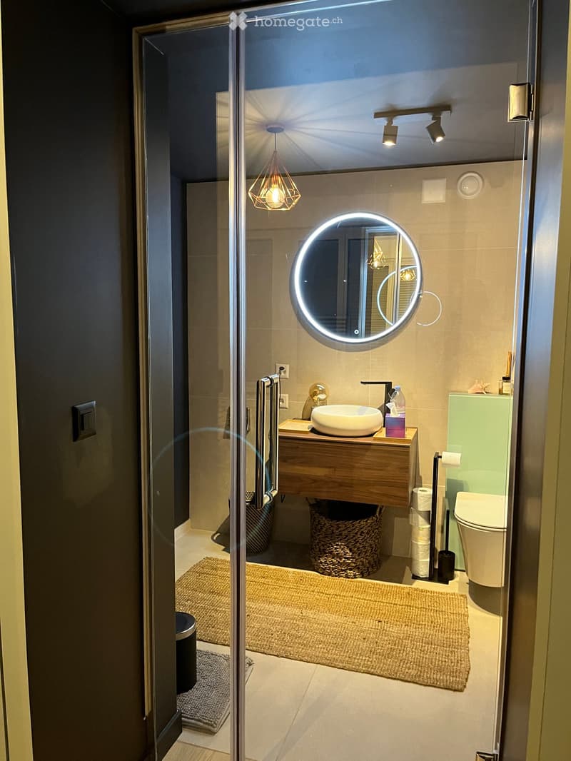 Modern bathroom with a floating wooden vanity, round illuminated mirror, toilet, and wicker basket on the floor.
