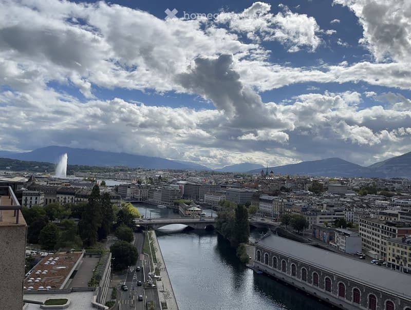 Panoramic city view from a balcony, showing a river, bridges, buildings, and distant mountains under a partly cloudy sky.