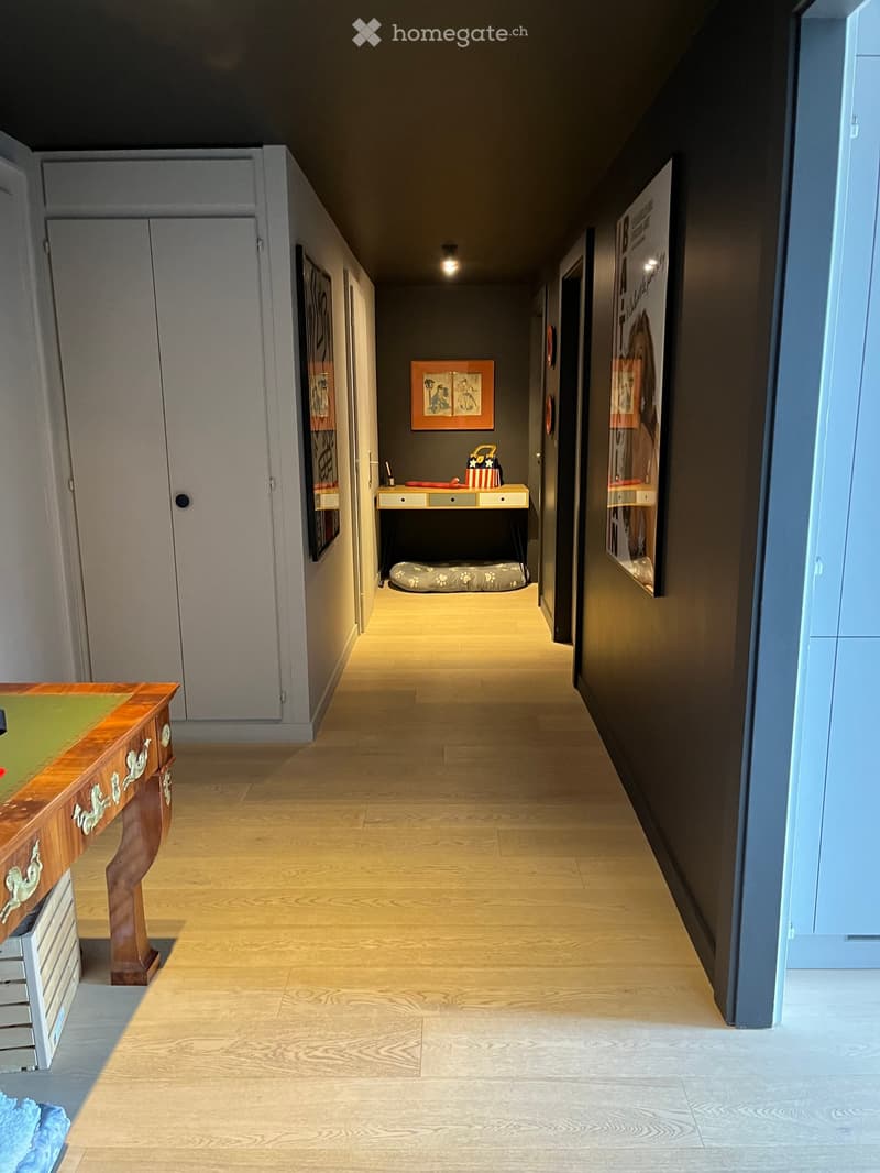 A modern hallway with light wooden flooring, dark walls, and several doors. There are framed artworks on the walls and a small table with a pet bed in the center.