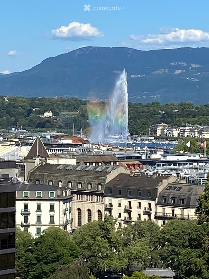 A panoramic view of a cityscape with a prominent fountain in the center, surrounded by historic buildings and a mountainous backdrop.