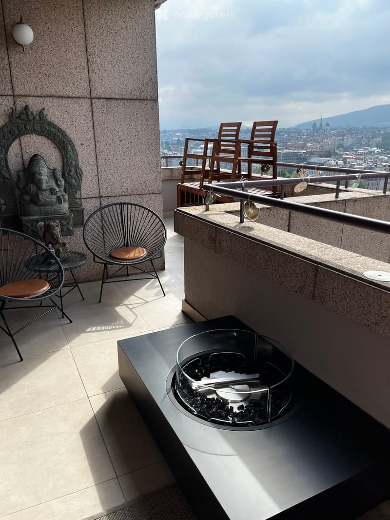 Balcony with a cityscape view, featuring a modern fireplace, two wooden chairs, and two metal chairs with brown cushions.