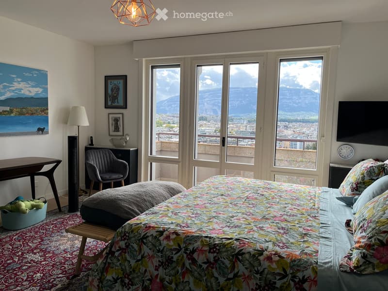 Bright bedroom with large windows offering a scenic mountain view, featuring a bed with floral bedding, a wooden stool, a grey armchair, a side table, a floor lamp, and decorative artwork.