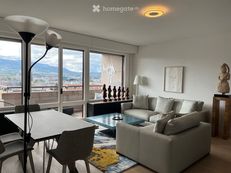 Modern living room with large windows offering a scenic view, a grey sectional sofa, a blue coffee table, a dining table with chairs, and decorative items including a statue and artwork.