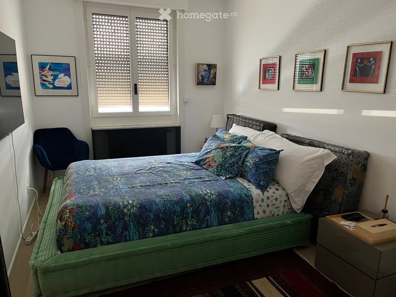A cozy bedroom with a double bed, nightstand, chair, and several framed artworks on the walls. The bed is covered with a blue and green patterned duvet and matching pillows.