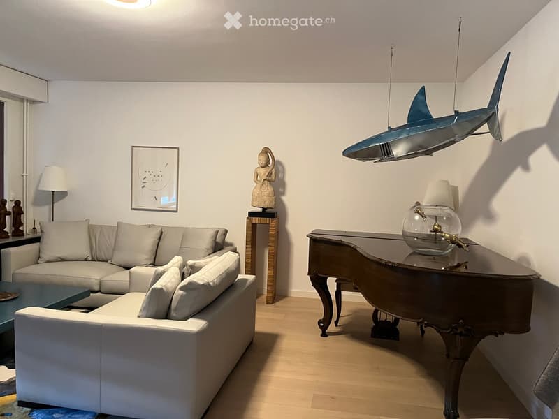 Modern living room with a white sectional sofa, coffee table, decorative table with a fishbowl, hanging shark sculpture, and a small statue on a stand.