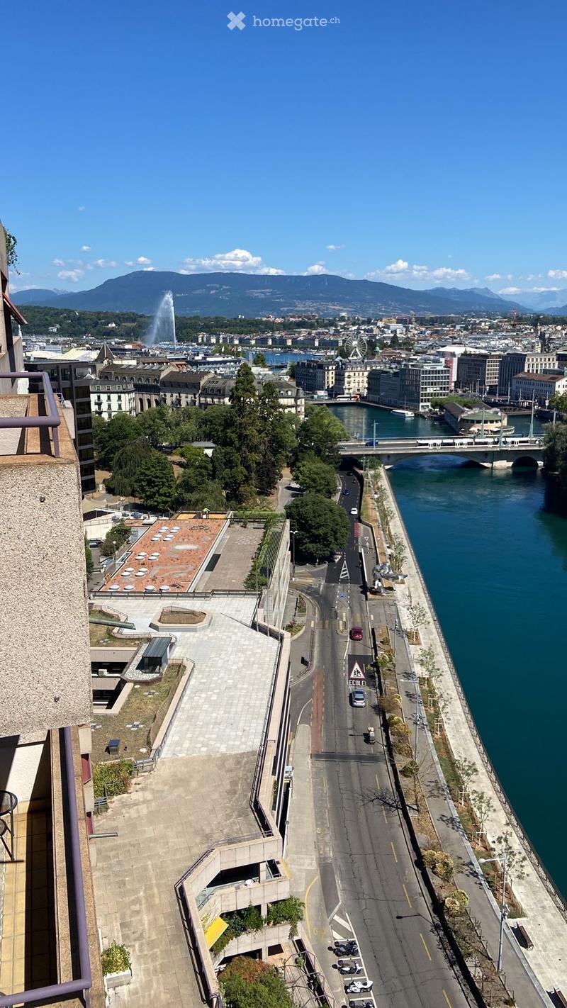 Aerial view of a cityscape with a river, bridge, and distant mountains, featuring a wide, empty road along the riverbank.