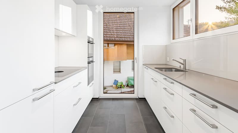 Kitchen. Modern white kitchen with sleek cabinets, stainless steel sink, and oven. Large windows provide natural light, and the floor is dark grey tile.