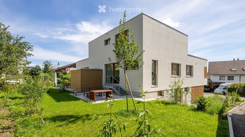 House. Modern two-story house with white exterior, large windows, and a small garden area with a wooden table and chairs.