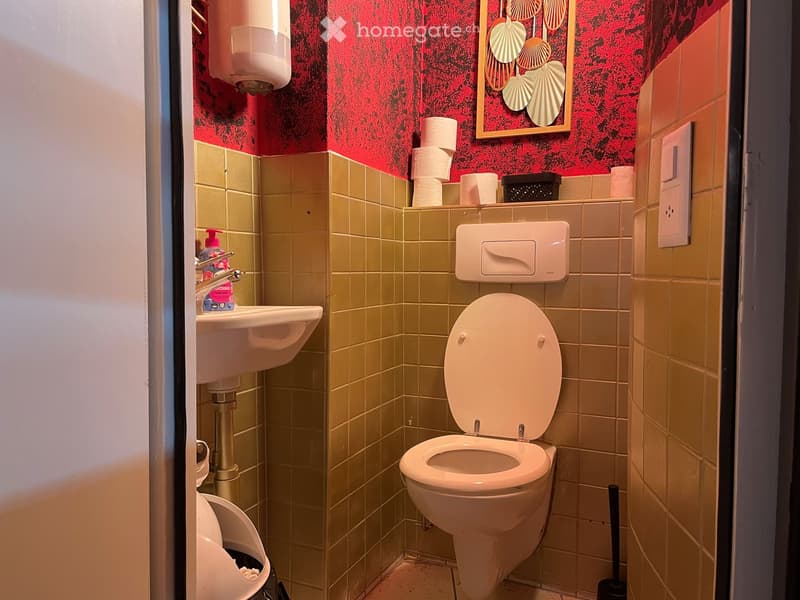 Small bathroom with beige tiled walls and floor, a toilet, a sink with a faucet, toilet paper, and a hand sanitizer dispenser on the wall.