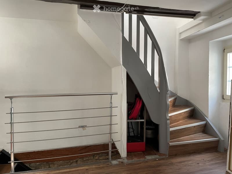 Modern hallway with wooden flooring, white walls, and a staircase with wooden steps and a metal railing. A small open closet with a red bag and other items is visible behind the staircase.