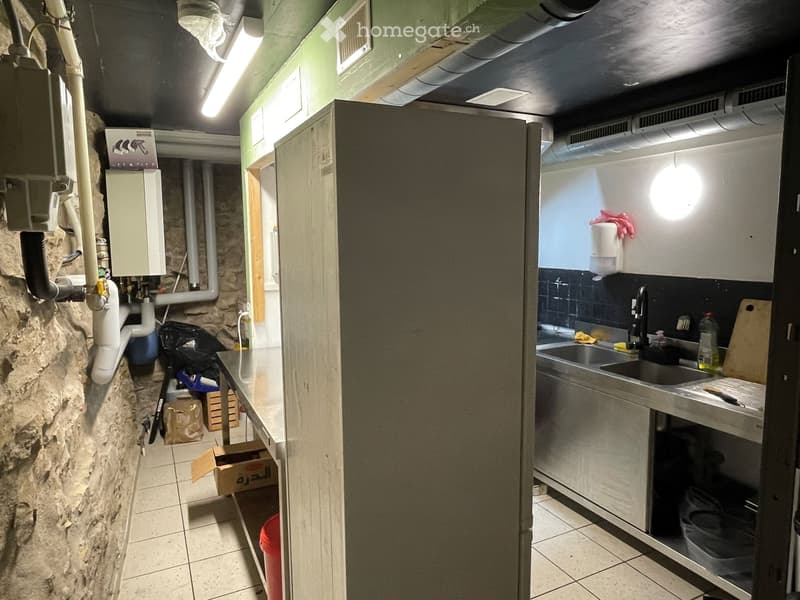 A compact kitchen with a stone wall, tiled floor, and stainless steel sink. Visible items include a faucet, cutting board, hand sanitizer, and a light fixture.