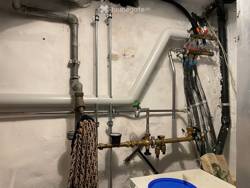 A utility room with exposed pipes, valves, and a towel hanging on the wall. There is a cup and a blue lid on a surface in the foreground.
