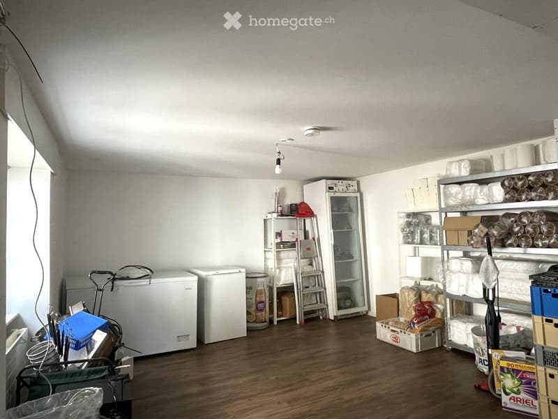 The room has wooden flooring, white walls, and is furnished with a refrigerator, shelves, a cart, and various boxes and items stacked around.