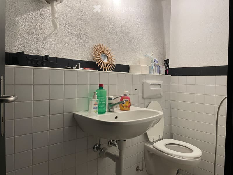 Bathroom with white tiled walls, a sink with a faucet, a toilet, and various toiletries on the sink counter.