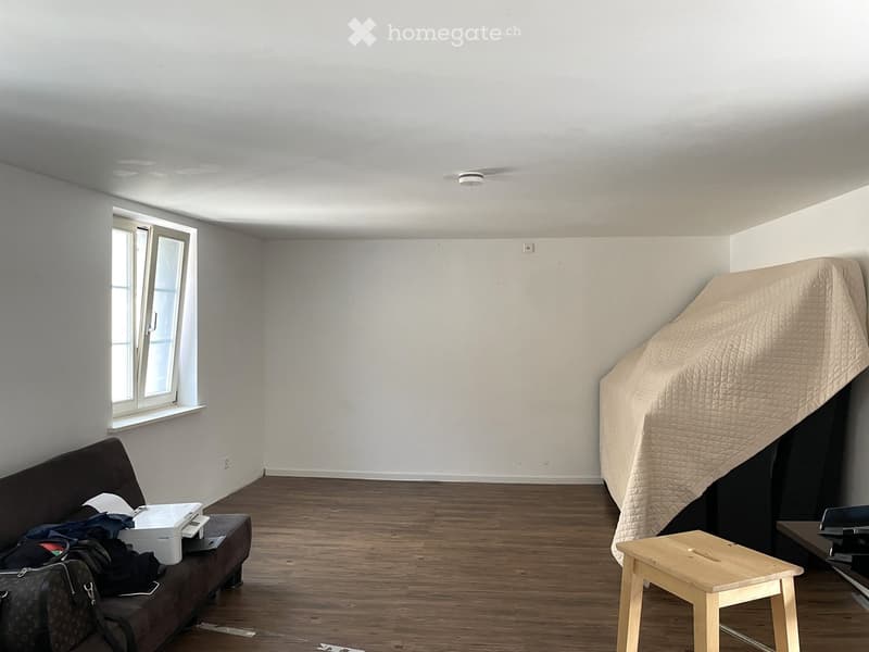 A furnished room with a dark brown sofa, a small wooden stool, a printer on the sofa, and a covered mattress on the floor.