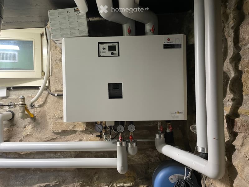 A wall-mounted water heating system with gauges, valves, and pipes against a stone wall.