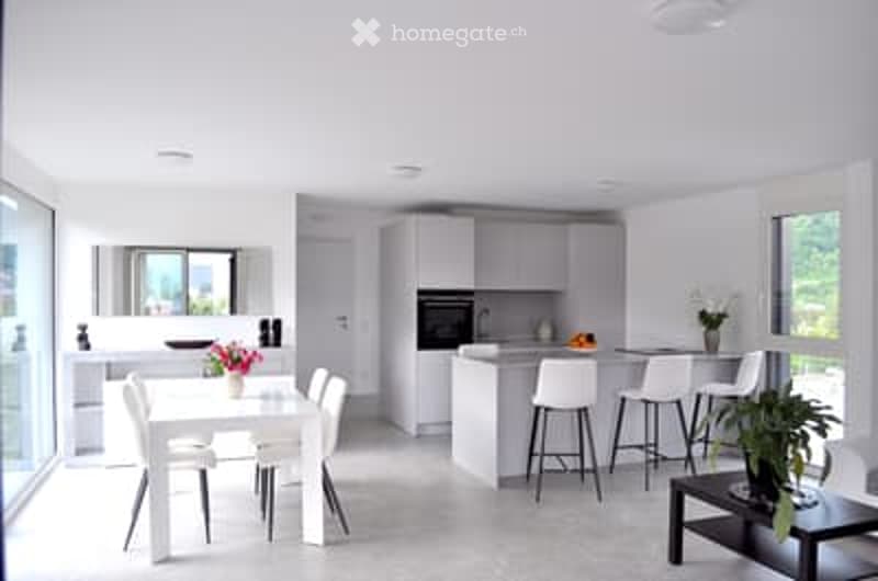 Bright, modern kitchen and dining area with white cabinetry, a central island, and a dining table with chairs. The space features large windows, a coffee table, and decorative plants.