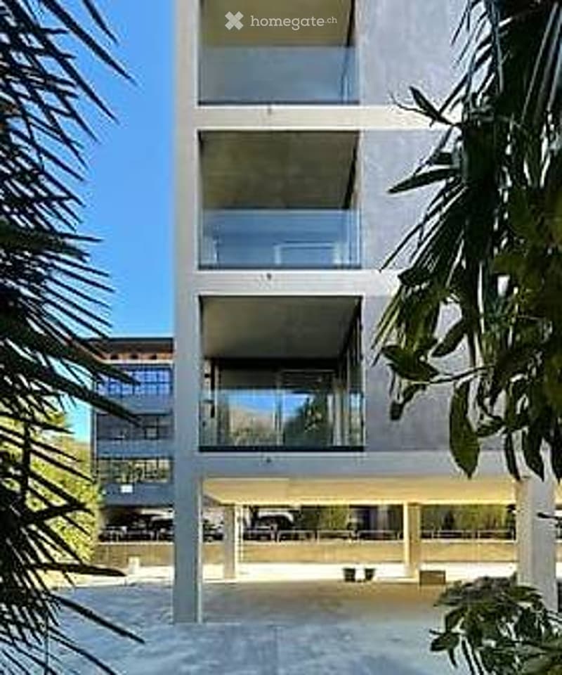 Modern multi-story building with large glass windows and balconies, surrounded by greenery and palm trees.