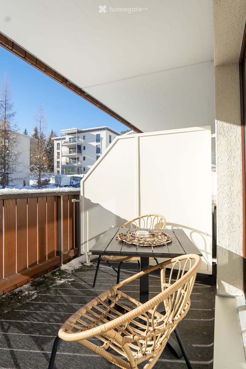 Outdoor wooden table and chairs, balcony wooden railing, white panels, and a view of buildings