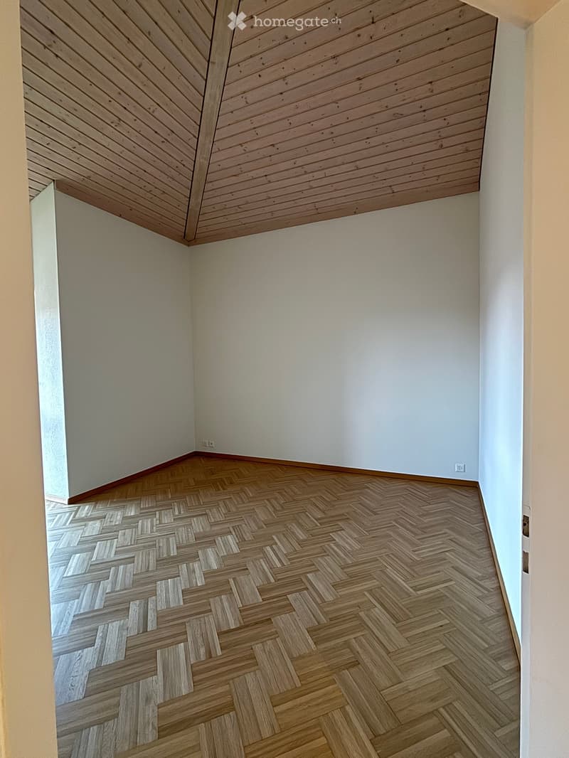 Empty room with herringbone-patterned wooden flooring and white walls, featuring a slanted wooden ceiling and visible electrical outlets.