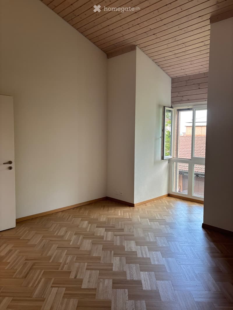 Empty room with wooden herringbone flooring, white walls, and large windows with wooden frames. The ceiling has exposed wooden beams.