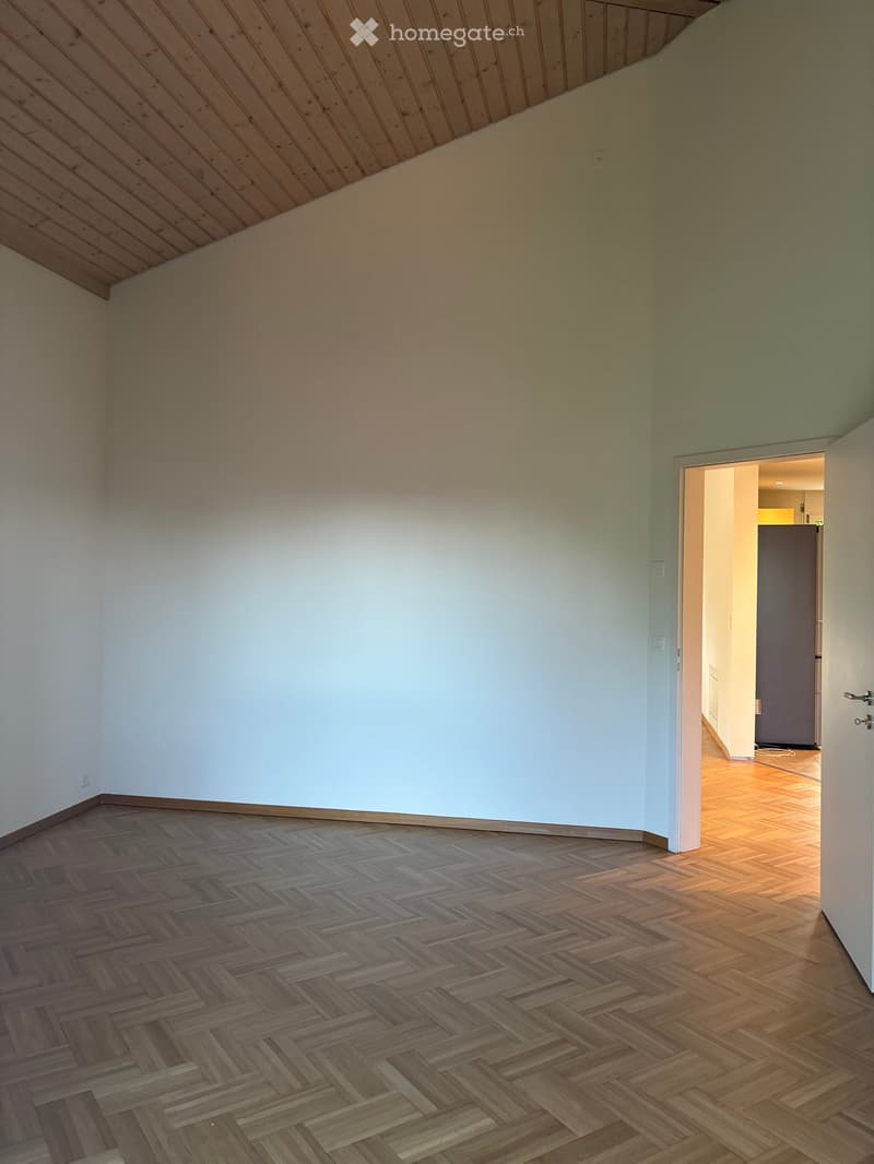 Empty room with wooden herringbone flooring and light-colored walls, featuring an open doorway leading to another room with wooden flooring and a dark-colored door.
