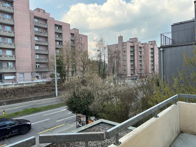 A view of a street with a car driving by, surrounded by trees and bushes, with a row of multi-story buildings in the background.
