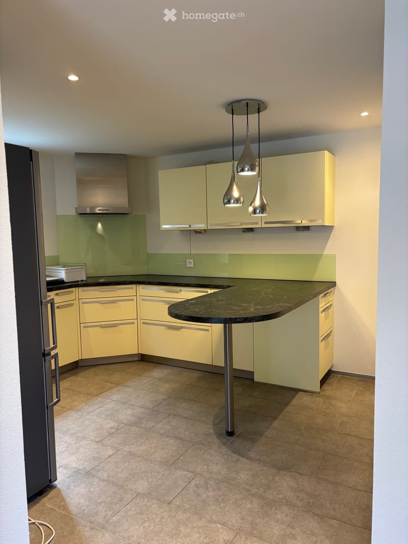 Modern kitchen with light yellow cabinets, a central island with a dark countertop, pendant lights, and a stainless steel range hood.