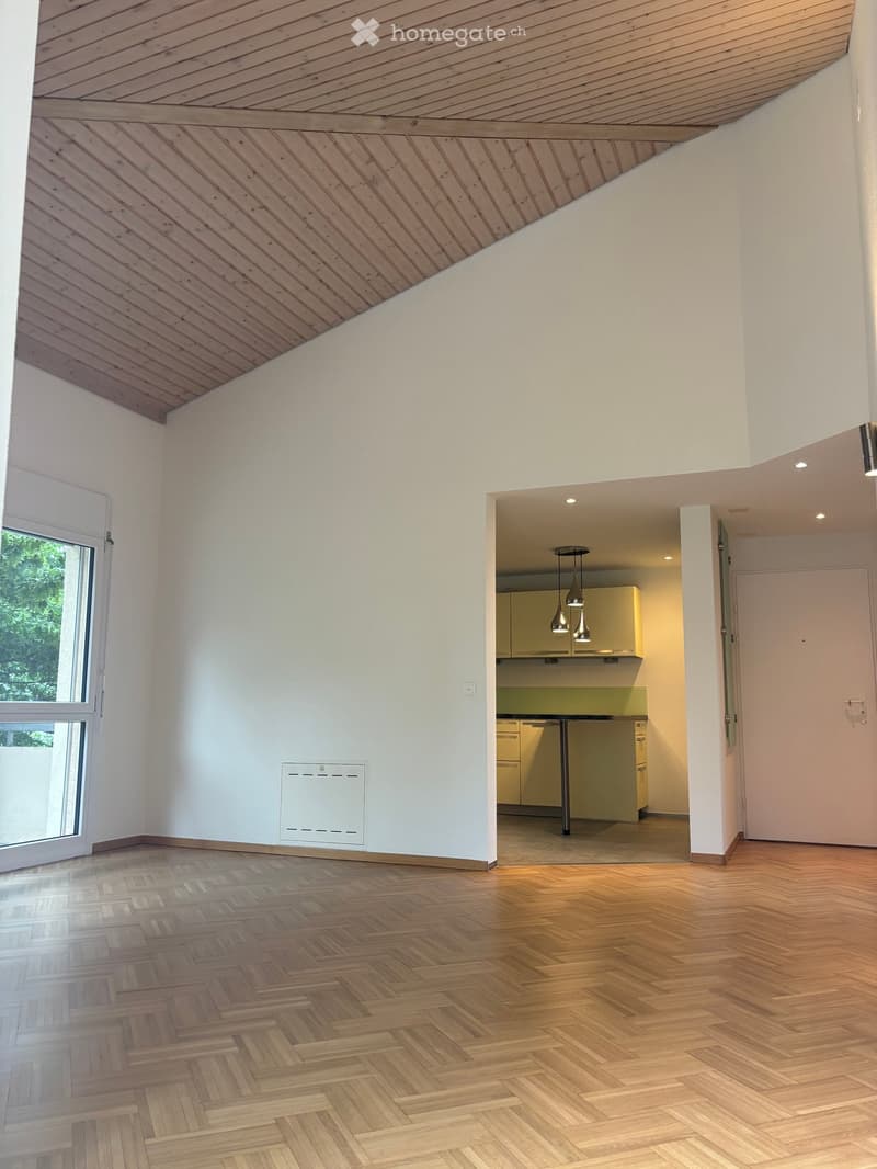 Empty room with wooden herringbone flooring, white walls, and a slanted wooden ceiling. A doorway leads to a kitchen area with cabinets and a table.