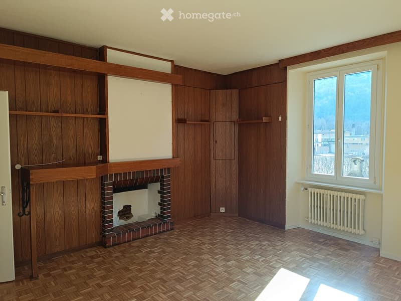 Empty room with wooden walls, herringbone wooden flooring, fireplace, radiator, and large windows offering a view of the outside.