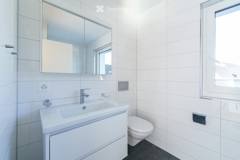 Modern bathroom with white tiles, a sink with a modern faucet, a large mirror, a toilet, and a small window letting in natural light.