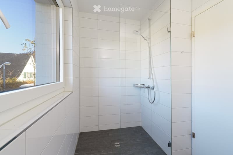 Modern bathroom with white tiled walls, dark grey tiled floor, glass shower enclosure, and a large window offering a view of the outside.