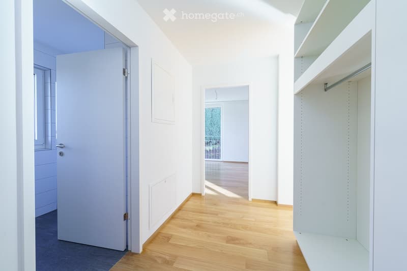 Empty room with white walls, wooden flooring, and built-in white shelving unit. The room features a closed door and a view of another room through an open doorway.