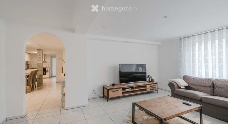 Furnished Room. Modern living room with a grey sofa, wooden coffee table, flat-screen TV on a wooden stand, and a dining area visible in the background.