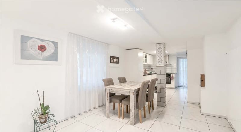 Furnished Room. Bright, modern dining area with a wooden dining table and four chairs, adjacent to a compact kitchen with white cabinets, an oven, and a sink. The space features white walls, tiled flooring, a large window with a sheer curtain, and decorat