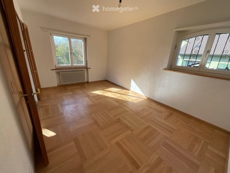 Empty room with wooden flooring, white walls, and two windows with metal grilles. A radiator is visible under one window, and a door is open on the left side.