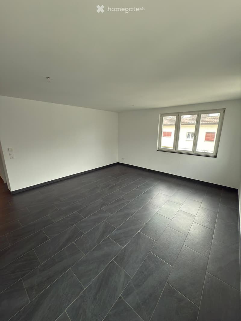 Empty room with light grey walls, dark grey tiled floor, and large windows showing a view of neighboring buildings.