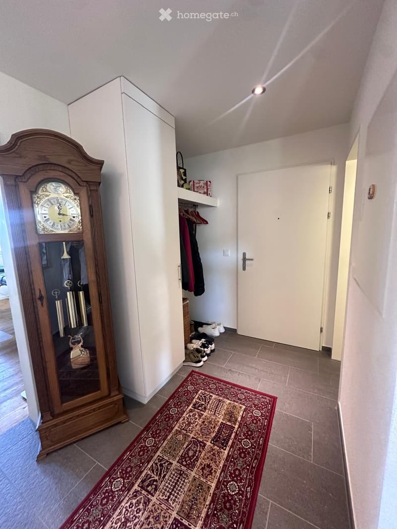Hallway with tiled floor, large wooden grandfather clock, small shelf with coats and shoes, and a patterned red runner rug.