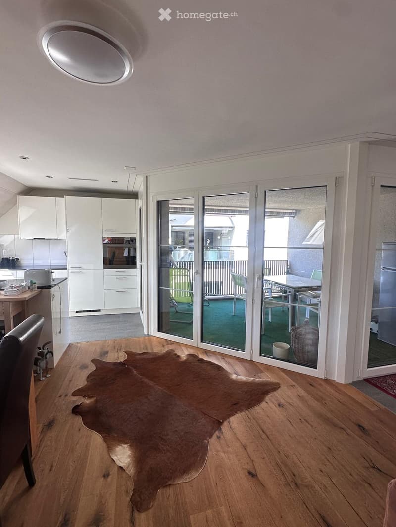 A modern living room with wooden flooring, a large cowhide rug, a chair, and a view through large glass doors to a balcony area with outdoor furniture.